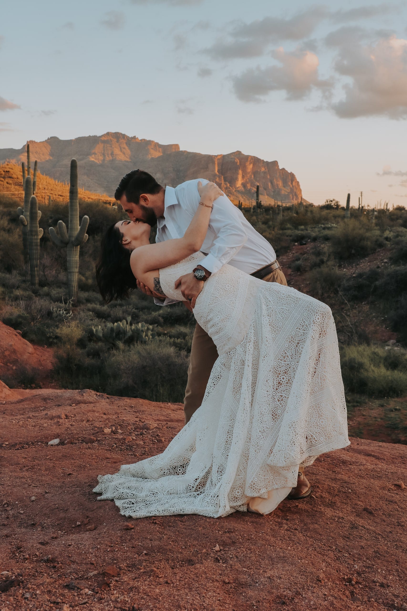 Groom dips his bride on red desert rock with the Superstition Mountains and saguaros behind them